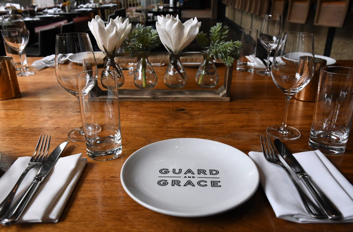 A wooden dining table set with white plates, glasses, cutlery, and a floral centerpiece. The plate has "Guard and Grace" printed on it.