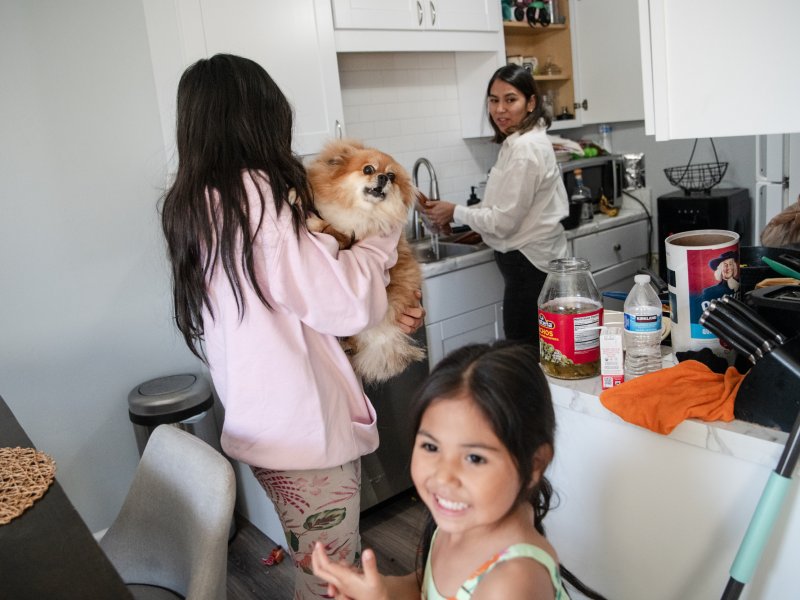 A woman washes dishes in the kitchen as one daughter holds a dog and another smiles toward the camera