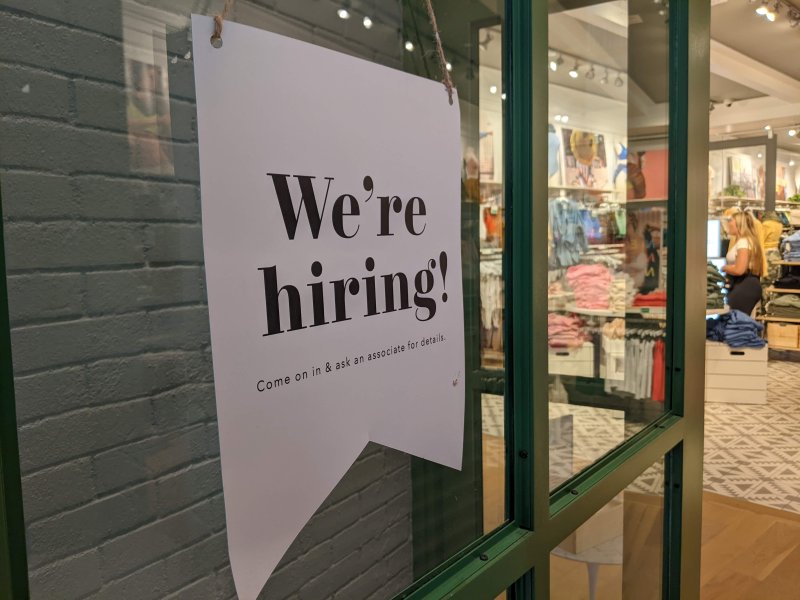 A "We're hiring!" sign is displayed on a glass door of a clothing store, with instructions to ask an associate for details visible. In addition to the exciting job opportunity, the store offers paid family leave. The store interior with clothes and other items is seen in the background.