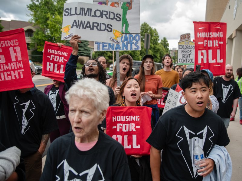 A group of people protest on a sidewalk, holding signs with messages like "Housing is a human right" and "Slumlords Awards." Several people wear black shirts with a white graphic.