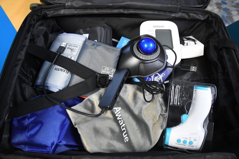 Open suitcase containing medical and electronic equipment, including a blood pressure monitor, a hand-held electronic device, and accessories such as cables and carrying cases.