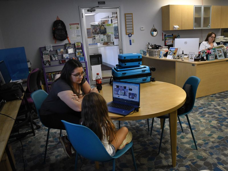 A woman and a child sit at a round table with a laptop in a library, while another woman works at a desk in the background.