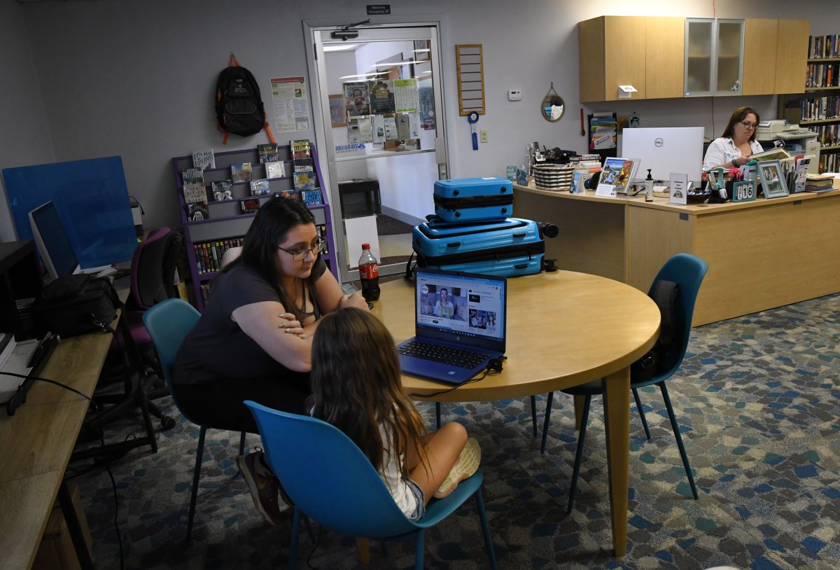 A woman and a child sit at a round table with a laptop in a library, while another woman works at a desk in the background.