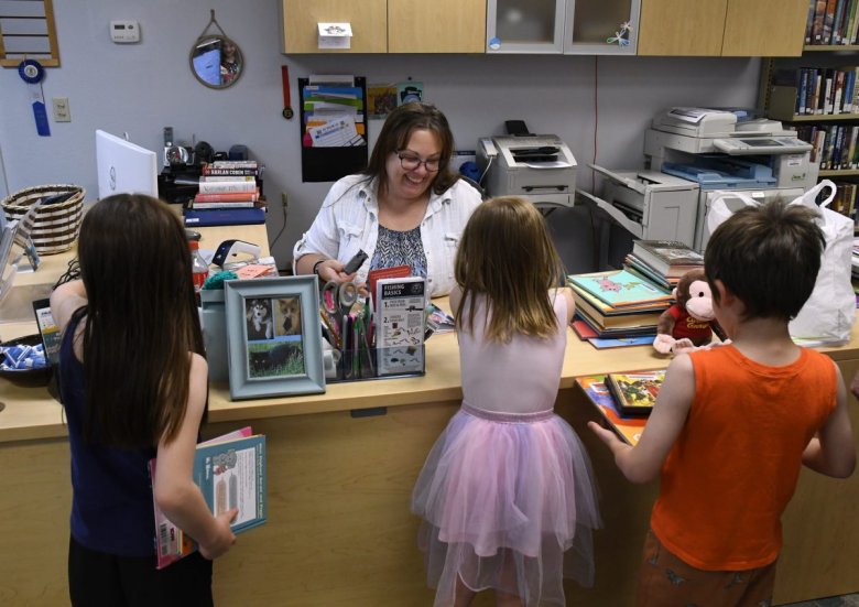 A librarian assists three children at the checkout counter of a library. The children are holding books, and various office supplies and equipment are visible on and around the desk.