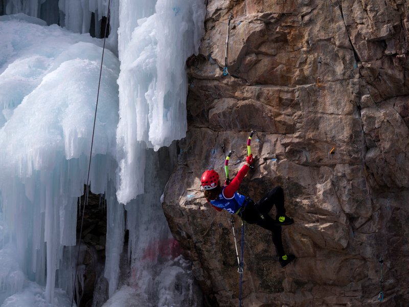 climber ascends on a large brown rock wall with an ice wall on the left side