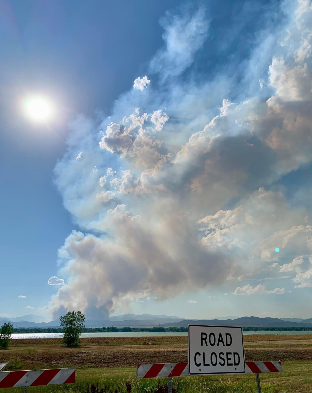 Smoke billows from mountains in the distance, beyond a lake and a sign reading road closed