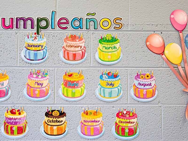 A classroom wall display shows birthday cakes representing each month of the year from January to December, with the word "Cumpleaños" above them. Balloons in various colors are attached to the right.