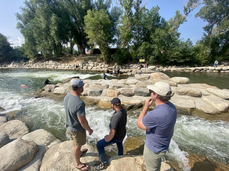 Three men stand on rocks, observing a river with rapids, surrounded by trees and other people. One man wears shorts, and the others wear jeans.