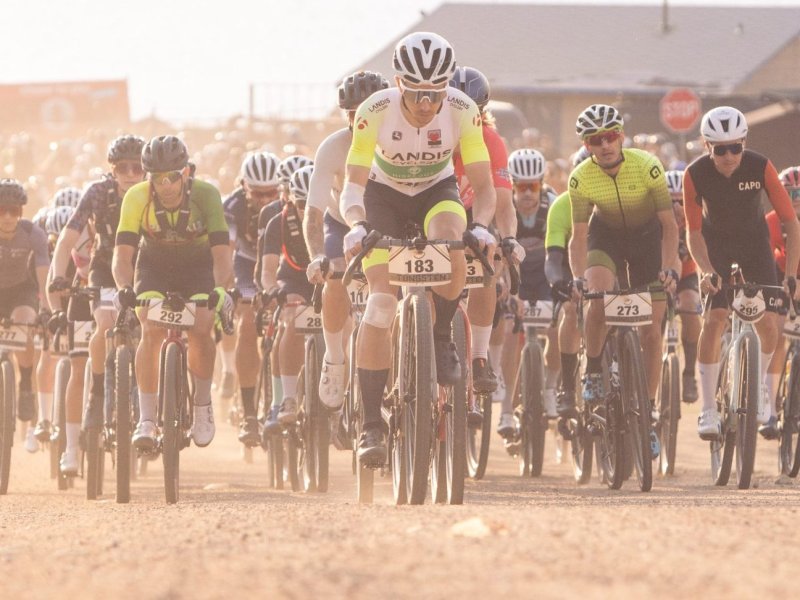 A group of cyclists ride on a dusty road during a race, with competitors wearing numbered jerseys and helmets. Buildings and parked cars are visible in the background.