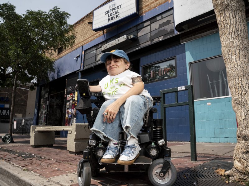 A person sitting in a motorized wheelchair on a sidewalk in front of a blue-brick building with a sign that reads "Support Local Music." The person is wearing a cap, t-shirt, and jeans.