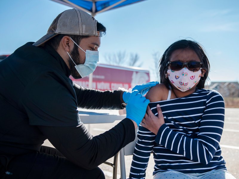 A healthcare worker wearing gloves and a mask administers a COVID vaccine to a child with glasses and a mask, outside in a tent-covered area. The child is in a striped shirt and is looking away.