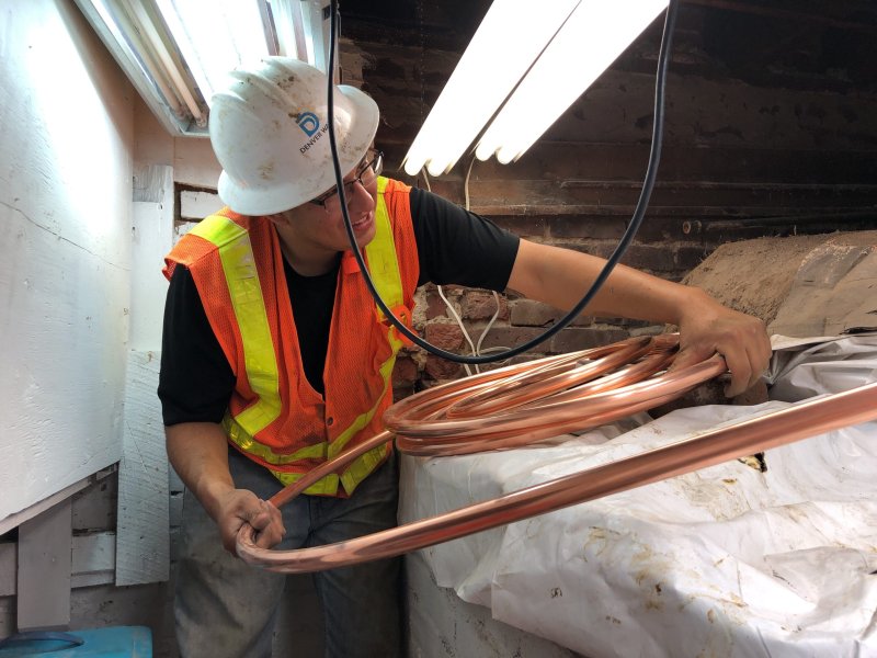 A man in a Denver Water hard hat and construction vest wrangles pipes inside a cramped space.