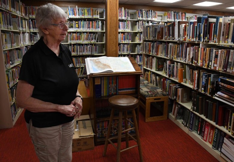 An elderly woman is standing next to a high table with an open book in a library, surrounded by shelves filled with books.