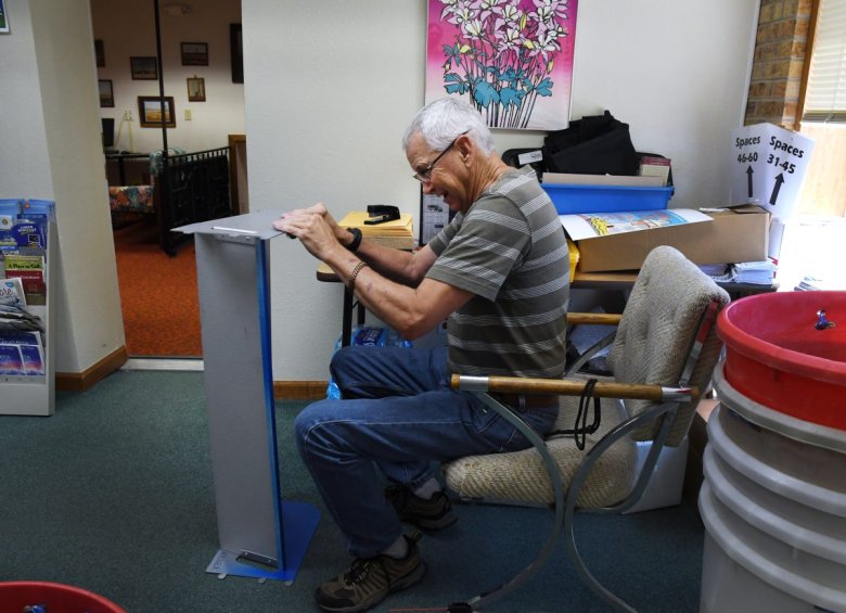 An elderly man wearing a striped shirt is seated in a chair and appears to be assembling a metal structure indoors. Various tools and materials are visible around him.