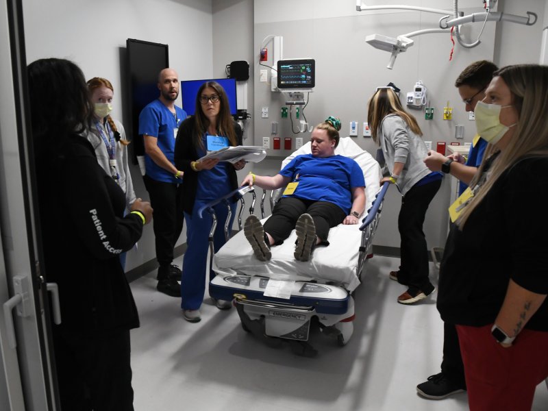 A group of medical professionals gathers around a nurse laying on a hospital bed in a medical room. The nurse is wearing a blue top, and some staff members are wearing masks and blue uniforms.