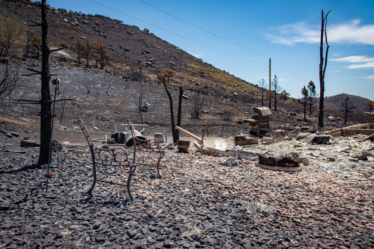 Burnt landscape with charred trees and remnants of metal furniture on the ground after the Stone Canyon fire. The area shows significant fire damage with a barren, scorched hillside in the background under a clear blue sky.