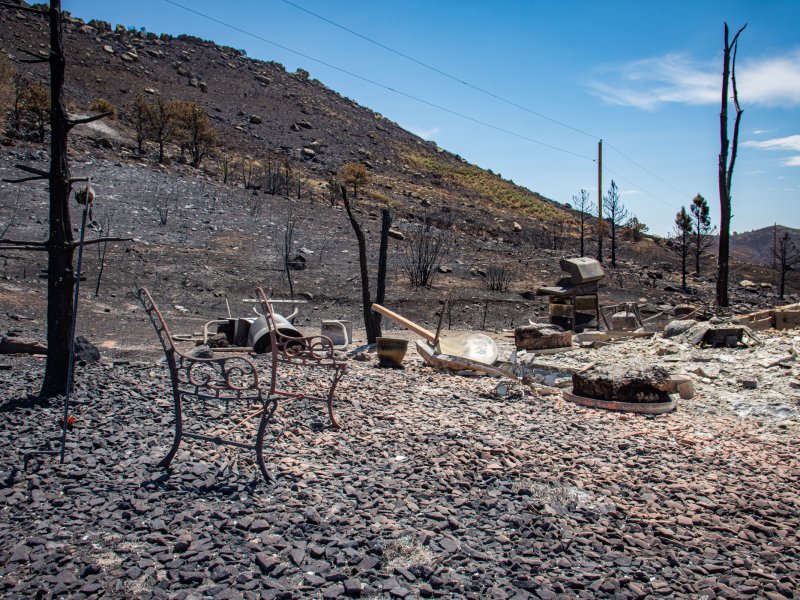 Burnt landscape with charred trees and remnants of metal furniture on the ground after the Stone Canyon fire. The area shows significant fire damage with a barren, scorched hillside in the background under a clear blue sky.