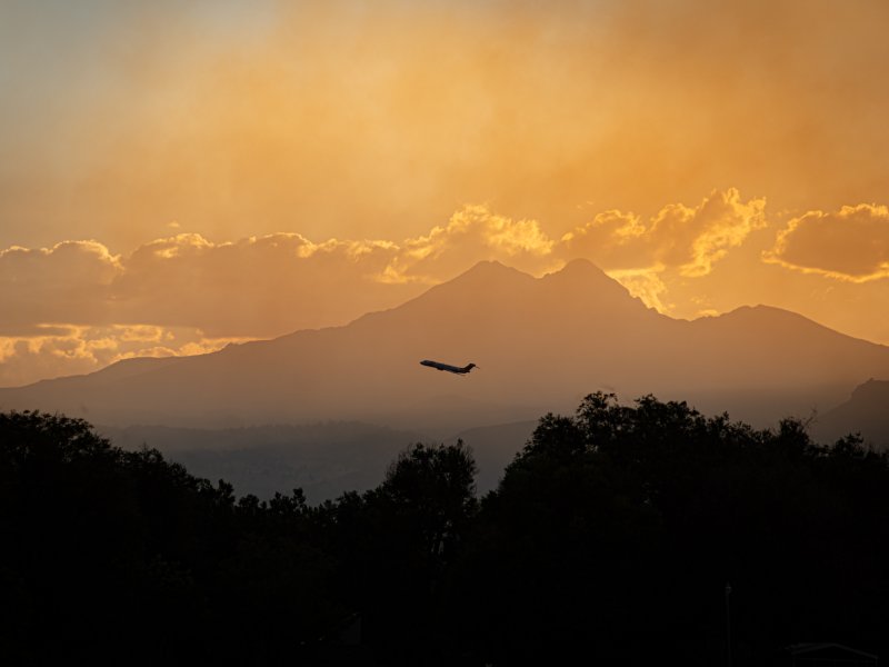 Silhouette of an airplane flying against a backdrop of Longs Peak and Mount Meeker and a glowing sunset sky with scattered clouds. Trees are visible in the foreground.