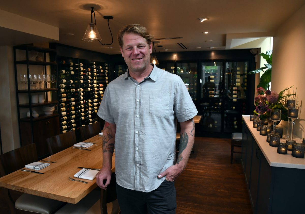 Alex Seidel stands smiling in a modern restaurant with wooden tables, wine racks, and candle decorations.