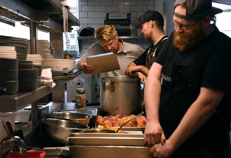 Three men work in a commercial kitchen; one reads a clipboard, another stirs a pot, and the third prepares food by a sink filled with peaches.