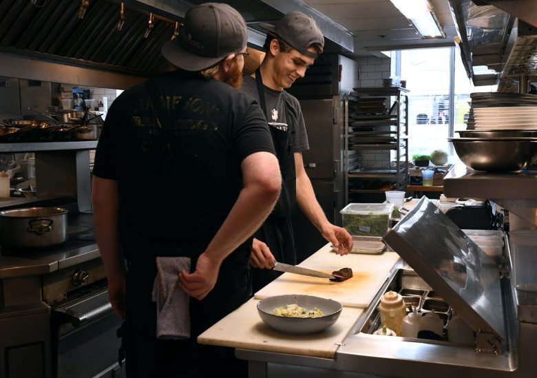 Two chefs in a restaurant kitchen preparing food; one chef is chopping ingredients on a cutting board while the other looks on with a towel over his shoulder.