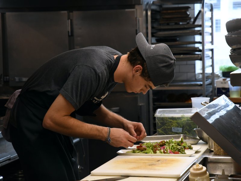 A person wearing a baseball cap and apron carefully prepares a salad in a commercial kitchen setting.