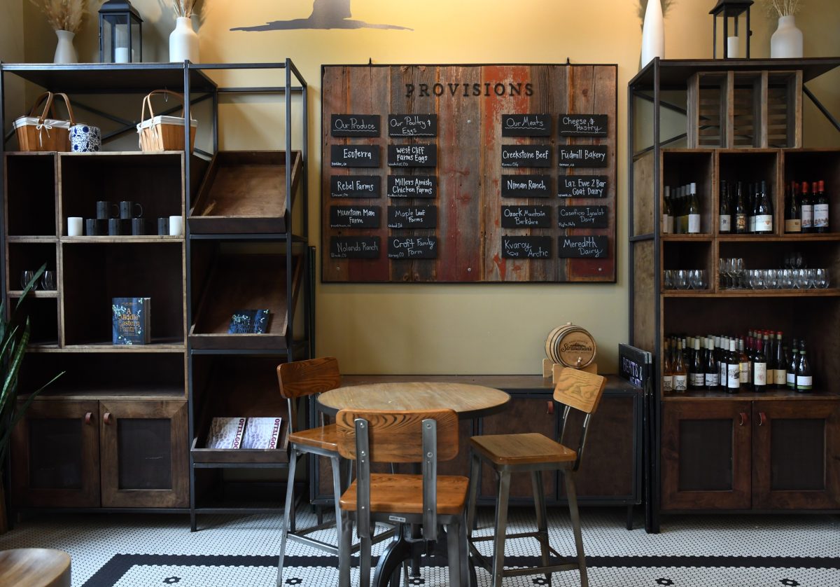 A cozy café interior with rustic shelves and a wooden menu board labeled "Provisions." The space includes a round table with three wooden stools surrounded by various displayed items and wine bottles.