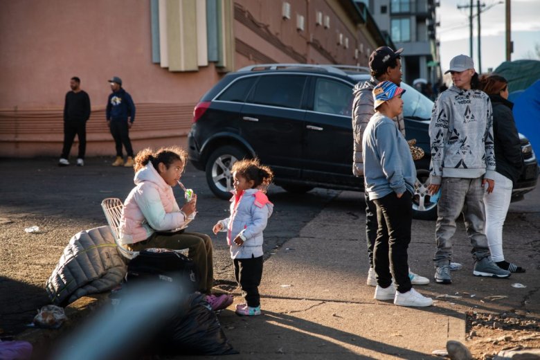 A young girl eats a snack while her little sister watches. Another group of people stand around talking on the side of the street.