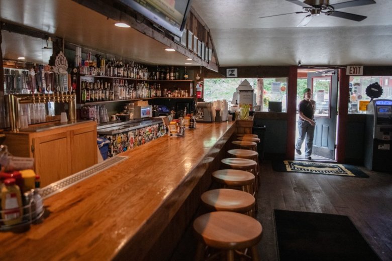 A man stands at an open door of a dimly lit bar with a long wooden counter and high stools. The bar is stocked with various bottles and beer taps.