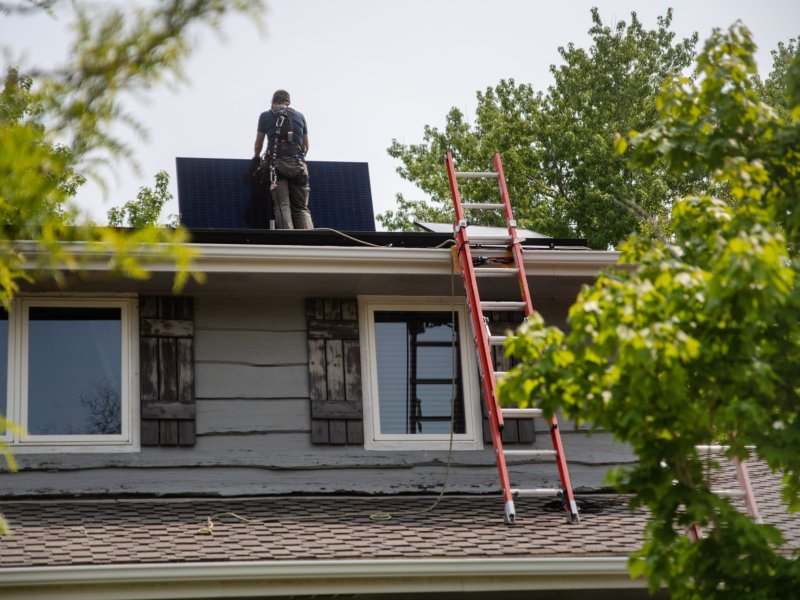 A worker installs solar panels on the roof of a residential home, accompanied by a red ladder extending from the ground to the roof.