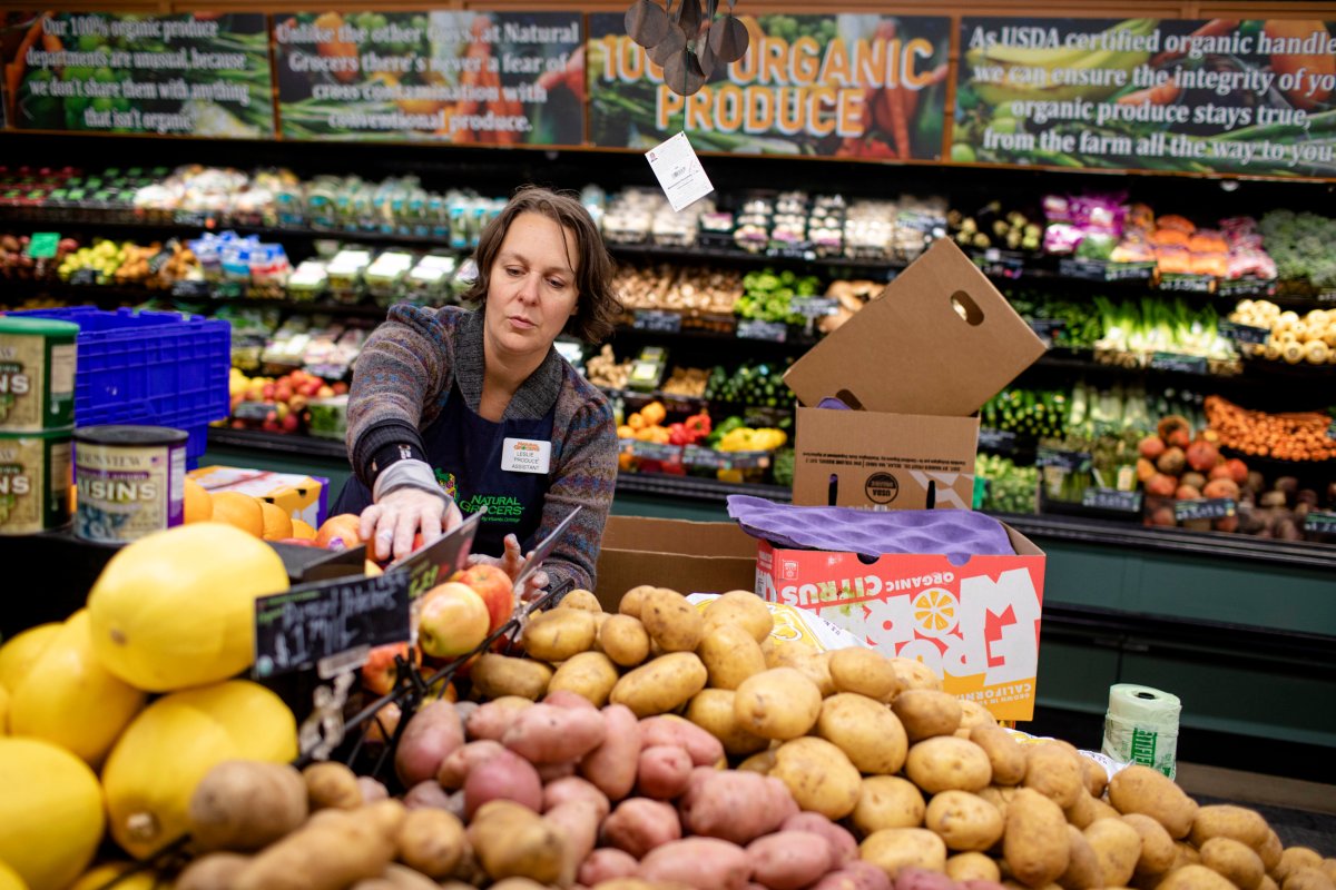 A grocery store employee arranges produce, including potatoes and fruits, in the organic section of a store filled with various fruits and vegetables.