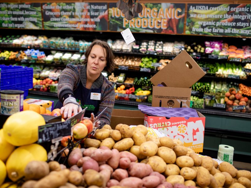 A grocery store employee arranges produce, including potatoes and fruits, in the organic section of a store filled with various fruits and vegetables.