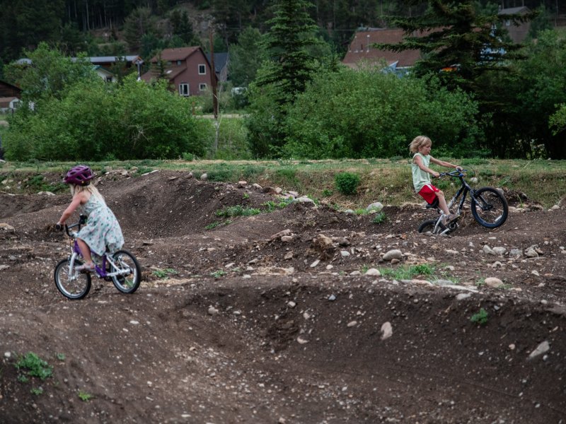 Two children ride bicycles on a dirt trail with small hills and curves. Trees, bushes, and houses are visible in the background.