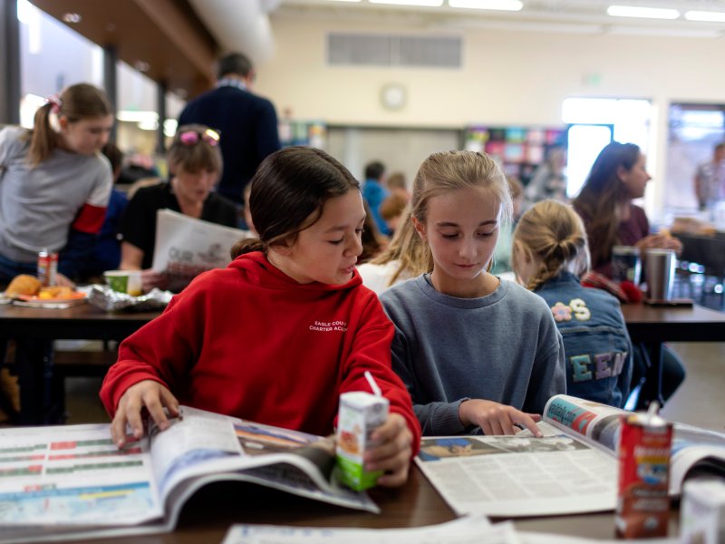 Young elementary school students sit at a table reading newspapers