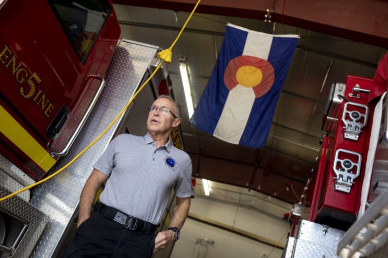 a man with polo shirt and glasses stands between to fire trucks inside a station with a colorado state flag in background