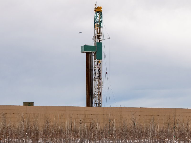 An oil drilling rig behind a concrete wall with sparse vegetation in the foreground under a cloudy sky.