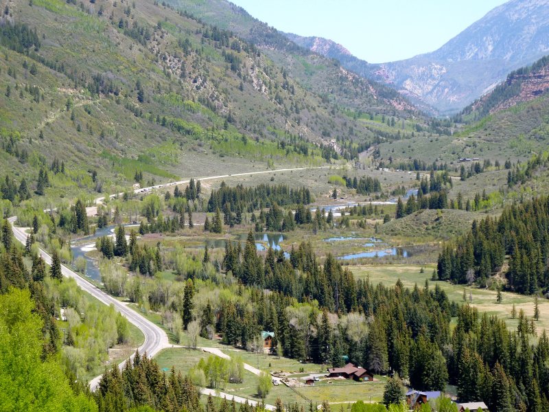 A scenic valley with green hills, a winding road, a river, scattered trees, and mountains in the background.