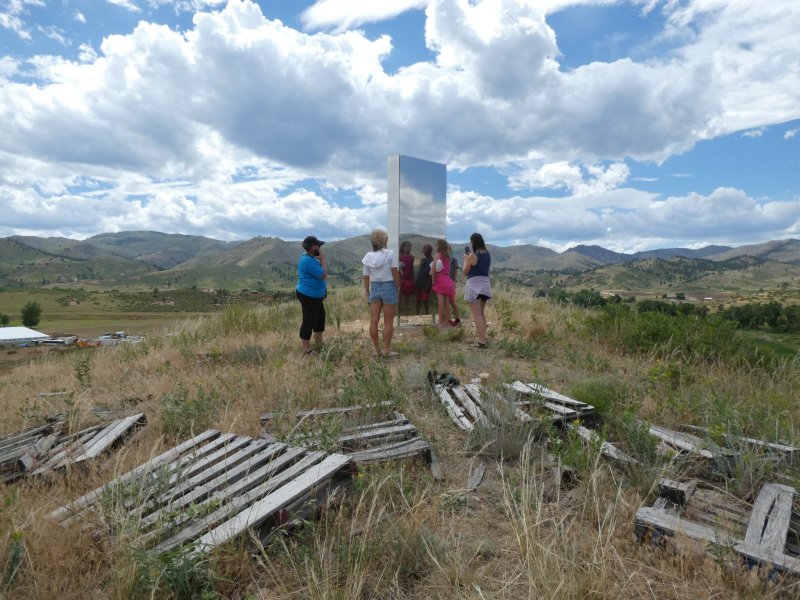 A group of people observe a tall metallic monolith on a grassy hillside with scattered wooden pallets, under a partly cloudy sky with mountains in the background.