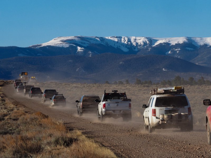 Cars and work trucks drive along a dirt road, including one loaded down with painting supplies