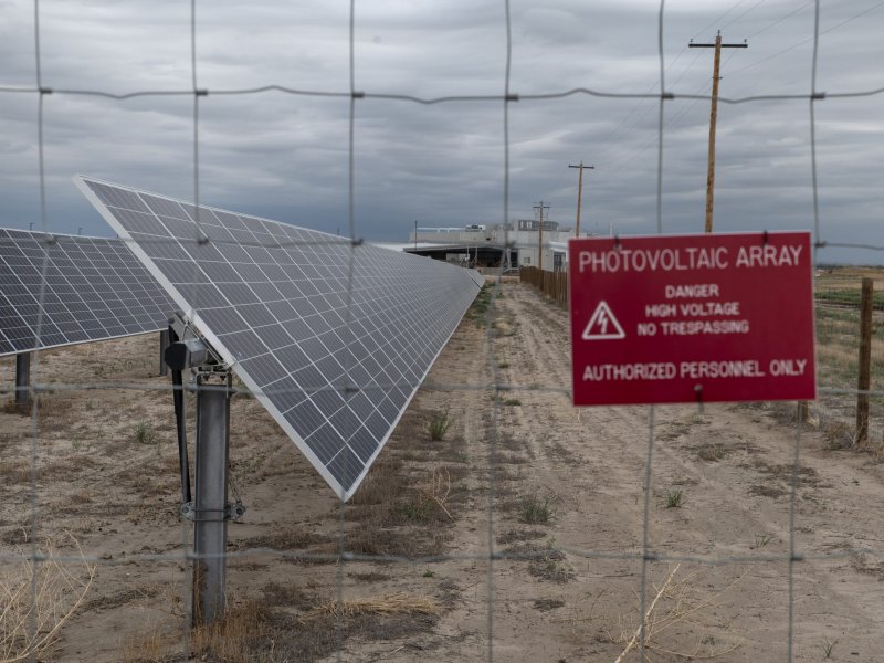 A fenced-off photovoltaic array with solar panels under a cloudy sky. A red sign on the fence reads, "Photovoltaic Array. Danger High Voltage. No Trespassing. Authorized Personnel Only.