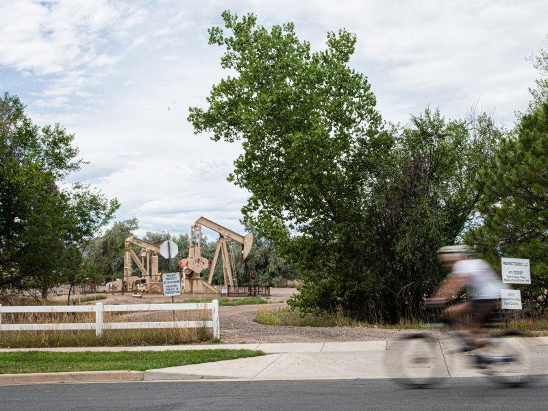 A photo of a fast-moving cyclist pasing the entrance to and oil and gas production site where idled beige pump jacks are visible just beyond the sidewalk of a neighborhood.