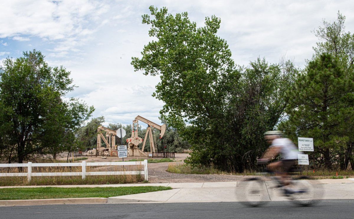 A photo of a fast-moving cyclist pasing the entrance to and oil and gas production site where idled beige pump jacks are visible just beyond the sidewalk of a neighborhood.