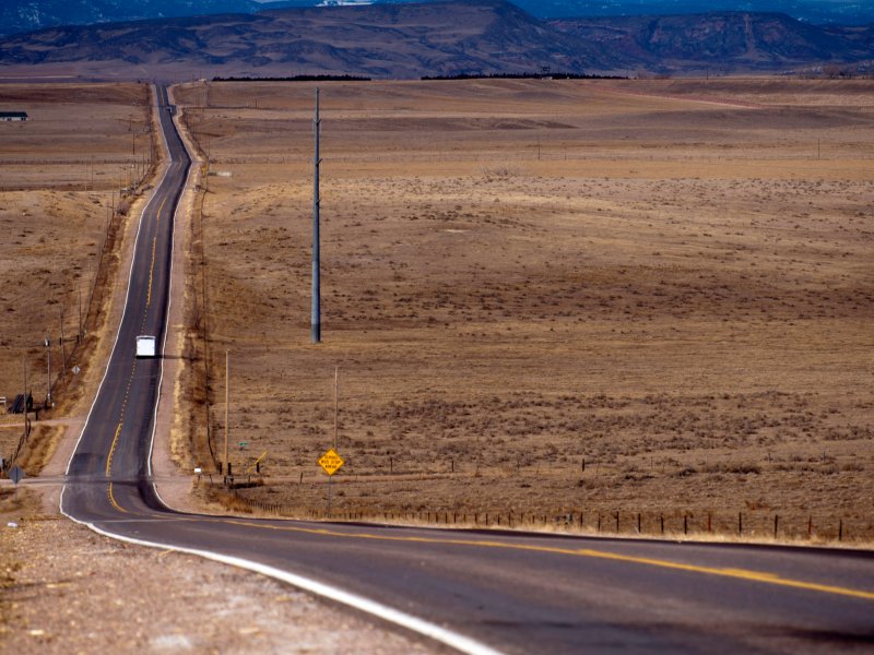 An empty, long, straight road stretches into the distance through a desolate desert landscape with mountains in the background. A single vehicle is driving on the road.