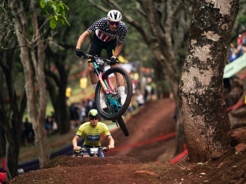 Cyclist Christopher Blevins in a star-spangled jersey performs a jump on a dirt trail through a forest, followed by another cyclist in a yellow jersey. Spectators are seen in the background.