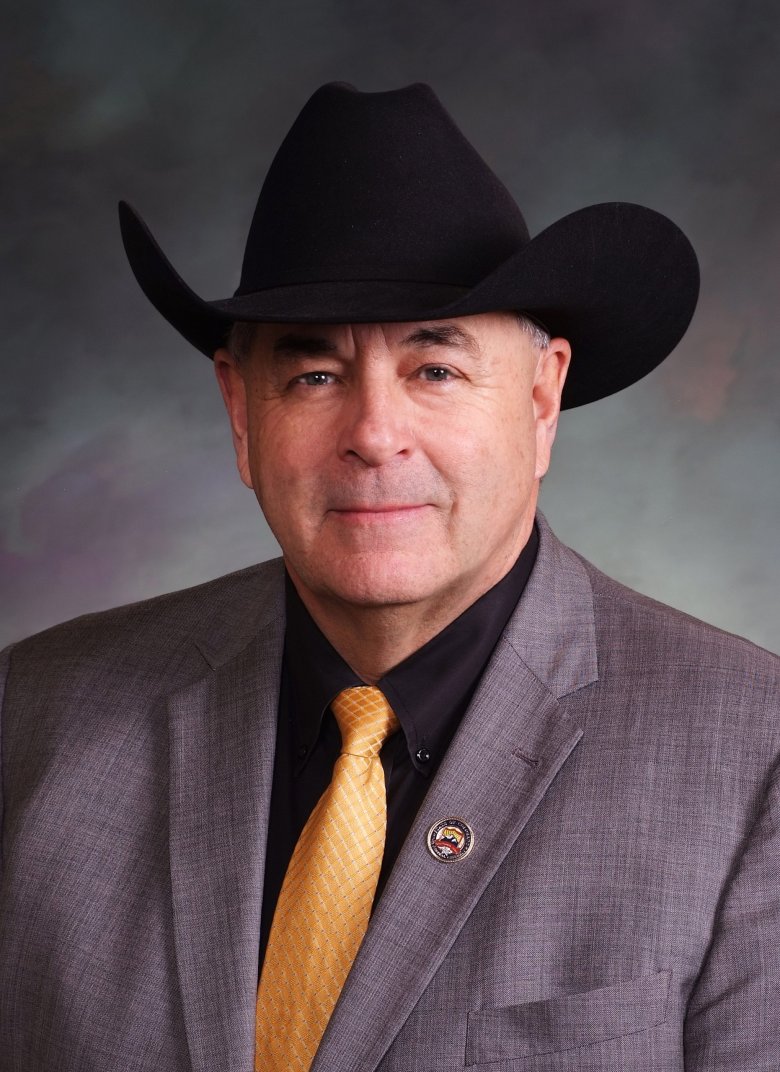 Cleave Simpson in a suit and yellow tie wearing a black cowboy hat, posing in front of a neutral backdrop.