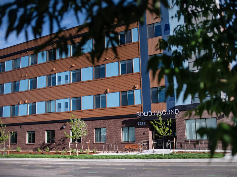 A four-story building with a mix of brown and blue colors, large windows, and the sign "SOLID GROUND" near the main entrance. Trees are in the foreground.