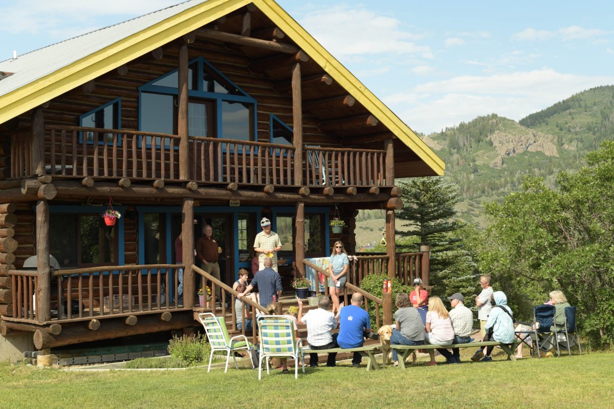 A group of people sit outside a large wooden cabin having a gathering. Some are seated on chairs in the grass, while others stand on the porch. Scenic mountains are visible in the background.