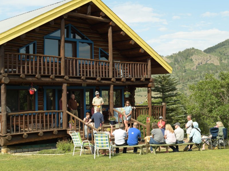 A group of people sit outside a large wooden cabin having a gathering. Some are seated on chairs in the grass, while others stand on the porch. Scenic mountains are visible in the background.