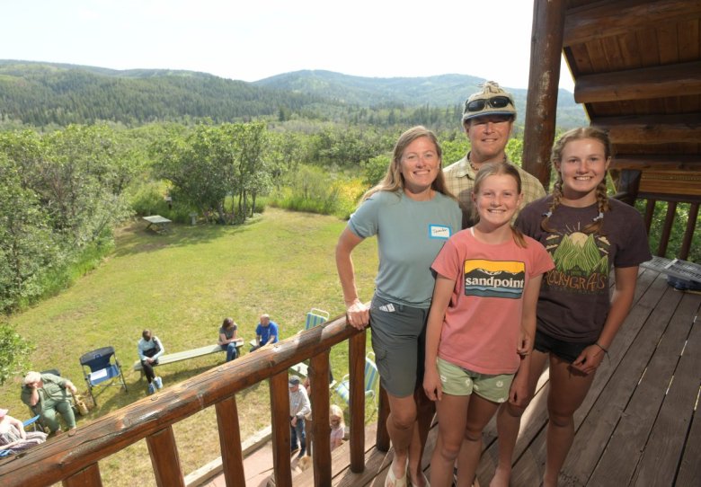 The Fernley family — a mom, dad and two daughters, pose for a picture on a wooden porch that overlooks a group of people mingling below.