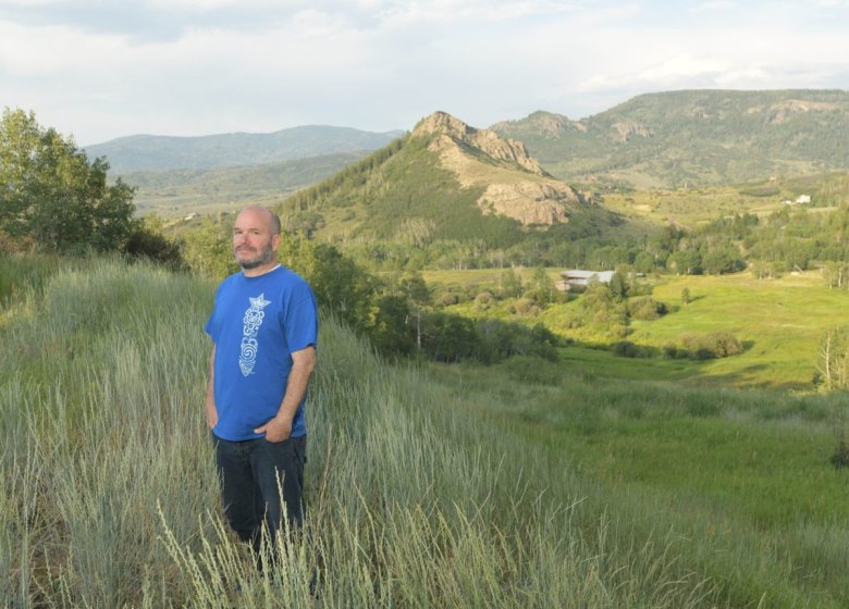 Josh Cook, wearing a blue shirt and black jeans, poses for a photo with his hands in his pockets while standing in long grass.